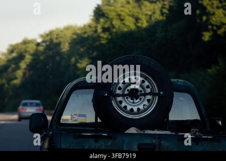 Close view of a weathered pickup carrying a spare tire on its tailgate while driving a tree lined road in soft evening light capturing a gritty mood o Stock Photo