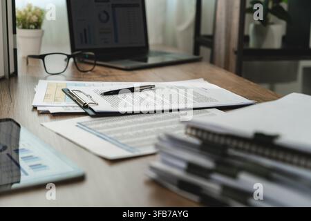 Printed business documents, clipboard, pen and eyeglasses arranged on an office desk beside a laptop, showing paperwork organization, report preparati Stock Photo
