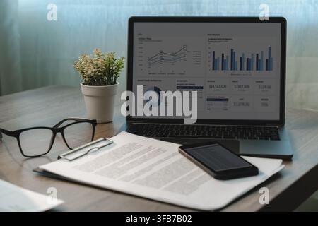 Laptop displaying business performance charts beside printed reports, smartphone and eyeglasses on an office desk, showing financial data analysis, re Stock Photo