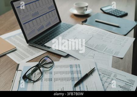 Laptop displaying financial dashboard beside printed spreadsheets, eyeglasses, pen, calculator on office desk, supporting document review, financial d Stock Photo