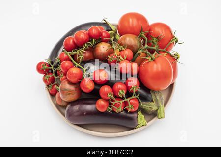 Different varieties of tomatoes and several eggplants are displayed on a plate, showing freshly picked seasonal vegetables from a home garden. Stock Photo