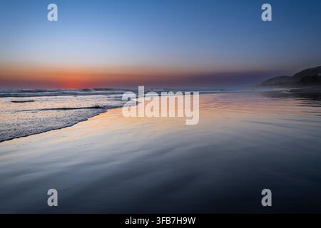 Gentle waves wash over wet sand at sunset, reflecting pastel hues along a tranquil Pacific shoreline. A distant headland fades into blue hour haze, ev Stock Photo