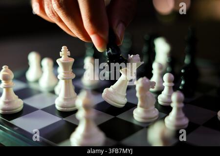 A closeup hand moving black knight among white chess piece on checkered board, creating strategic tension during intense indoor game with shallow dept Stock Photo