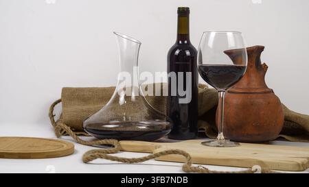 Rustic still life of red wine in a glass decanter and wine glass with an unlabeled bottle, aged clay jug, burlap, rope and wooden boards on white. Stock Photo