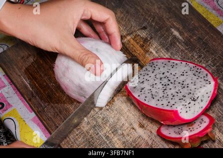 A dragon fruit slicing on wooden cutting board, hand holding fresh white flesh for food preparation and healthy eating at home Stock Photo
