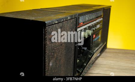 An angled perspective reveals a vintage black boombox resting on a wooden table against a vibrant yellow background, featuring an open cassette deck d Stock Photo