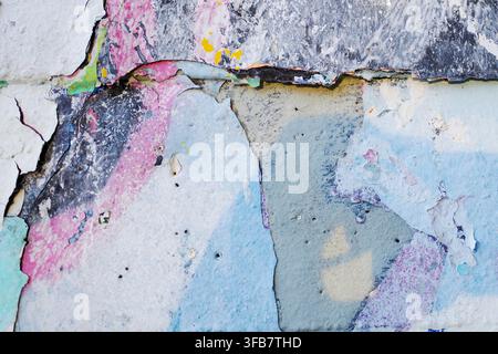 Peeling paint layers on a wall reveal a textured surface beneath. Multiple colors are visible, including blue, pink, black, and yellow from previous p Stock Photo