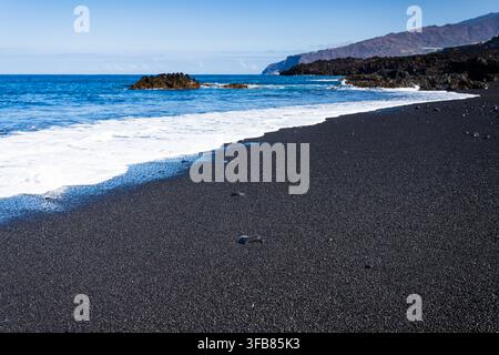 Vertical close-up perspective of white sea foam and gentle waves sweeping over smooth black volcanic sand along the ocean shore in La Palma. Stock Photo