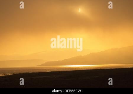 Golden solar eclipse over coastal mountains and water, casting a bright reflection across the sea under a dramatic hazy orange sky. Stock Photo