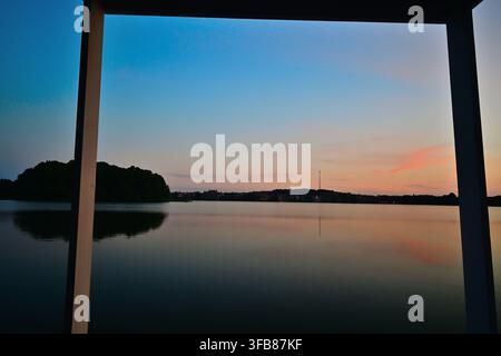 The tranquil reflection of the sunset on Lake Czos, framed by the pier pillars over the calm water at dusk, with the Mrągowo skyline in the distance. Stock Photo