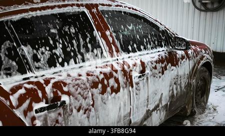 dirty car with foam on windows and body at car wash. soap bubbles covering the surface as part of cleaning process, washing procedure in progress, gri Stock Photo