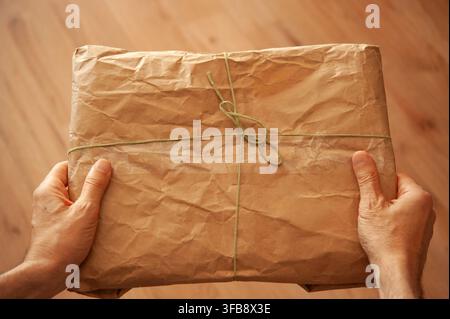 Hands hold a parcel wrapped in crumpled brown craft paper and tied with string over a warm wooden floor Stock Photo