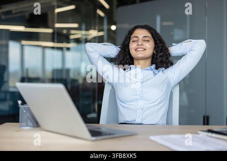 Young businesswoman leaning back in her office chair with arms behind head, smiling with closed eyes, finding relief and satisfaction after completing a task in a contemporary workspace Stock Photo