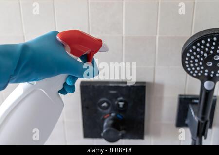 Close-up of a gloved hand holding a spray bottle and cleaning a shower faucet and tiled wall Stock Photo