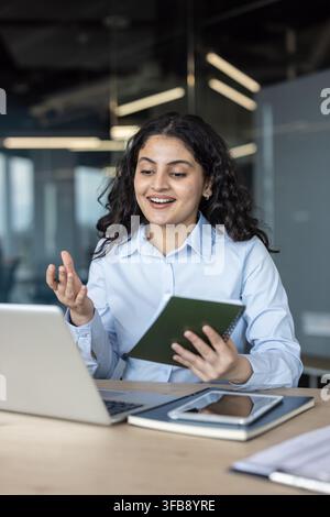 Young businesswoman participating in a video conference from her corporate office, actively discussing and taking notes while communicating through her laptop Stock Photo