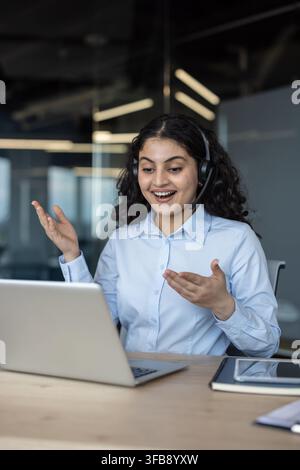 Young indian woman with headset smiling and gesturing during a video call, working on a laptop in a modern office setting, providing online support and consulting remotely Stock Photo