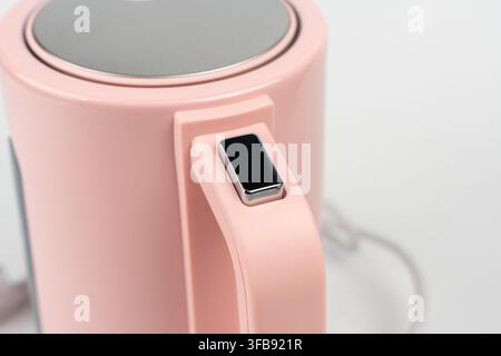 A macro shot highlights the handle of a pink electric kettle with its glossy black power button set into the grip, on a white background. Stock Photo