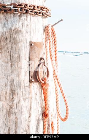 Rusty chain and orange mooring ropes hang from a weathered wooden pier beside calm water. Stock Photo