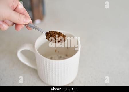 Spoon Releasing Instant Coffee Granules Into White Mug On Kitchen Counter With Hand Visible And Jar Blurred Behind In Neutral Interior Morning Beverage Preparation Closeup. Stock Photo