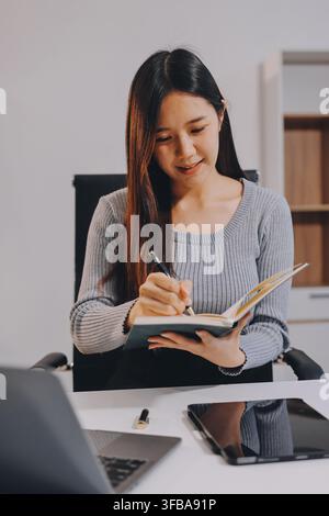 Young asian businesswoman is smiling and working on a laptop while sitting at her desk in a modern office Stock Photo