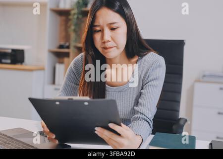 Young Asian woman sits at office desk rubbings her eyes and temples feeling pain. Stress. And exhaustion from long hours working on the computer Stock Photo