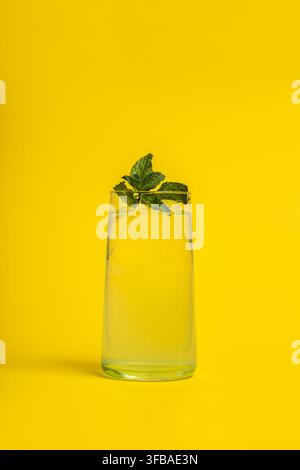 Clear glass of cold lemonade with ice cubes and mint leaves against a yellow background. The simple and fresh setup evokes summer energy, making it pe Stock Photo