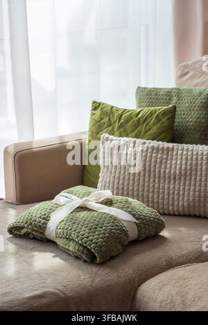 Soft textured green blanket neatly folded and tied with white ribbon positioned on sofa surface. Plush decorative pillows in green and beige tones cre Stock Photo