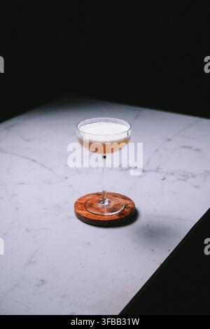 Whiskey sour with a thick foam top in a coupe glass on a wooden coaster on white marble against a dark background, drinks photography Stock Photo