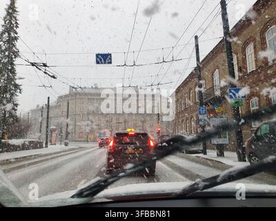 View from inside a car during heavy snowfall in an urban setting. Windshield wipers are visible in the foreground, actively clearing snow from the Stock Photo