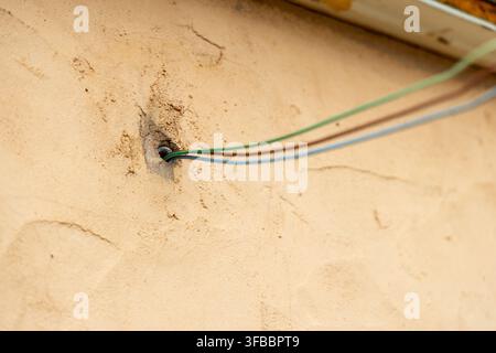 Three insulated electrical cables pass through a small opening in a beige, textured wall, suggesting construction or repair work Stock Photo