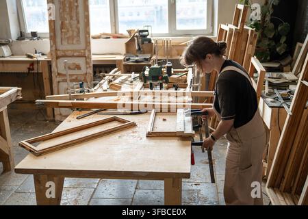 Woman crafter in restoration workshop fixating parts of vintage wooden window frame using clamps. Stock Photo