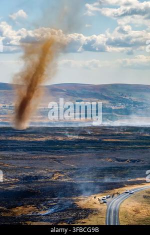 Tall vortex of smoke and dust rising from smouldering black ground after hill fire, spectators and vehicles gathered along rural road in summer Stock Photo