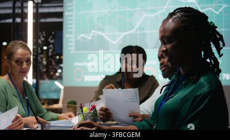 Diverse administrative team attending briefing conference at night to verify updates on regulatory changes or industry standards. Business professionals reviewing all data analysis. Camera A. Stock Photo