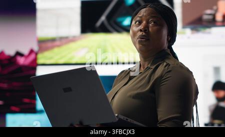 Woman uses laptop, enjoying leisure time with content on video streaming services. African american person watches media VOD broadcasts on notebook in front of digital backdrop with clips, camera B Stock Photo