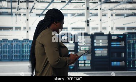Technician walking in data center with tablet, checking hardware for system stability. Worker overseeing server farm with device, configuring infrastructure rigs, camera B Stock Photo