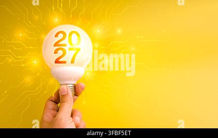 Hand holds glowing white globe light bulb showing new year 2027 against bright digital circuit background. Technology innovation, future planning, cre Stock Photo