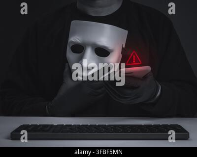 Dark conceptual image of masked cybercriminal scammer holding smartphone beside red warning icon and keyboard. Digital identity theft, phishing, fraud Stock Photo