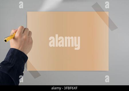 Close-up of a hand with a yellow pen beside a large blank paper poster taped to a gray wall, designed for copy space, notes, planning, advertising, ed Stock Photo