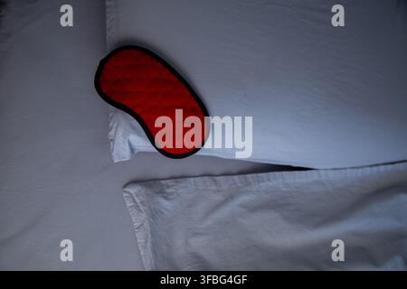 A close-up view from above of a red sleep mask lying on a pillow against the background of a bed Stock Photo