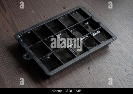 Top-down view of a black ice cube tray filled with water placed on a dark wooden table. The reflective liquid surface and scattered droplets highlight Stock Photo