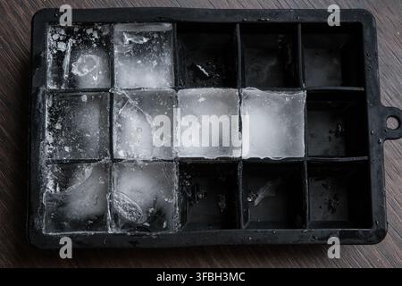 Top-down view of a black ice cube tray filled with icy water and frozen cubes on a wooden table. The frosty texture and empty compartments highlight t Stock Photo