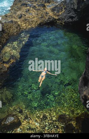 Woman in a blue bikini enjoying vacation, floating effortlessly on her back in a volcanic natural pool with clear turquoise water surrounded by dark r Stock Photo