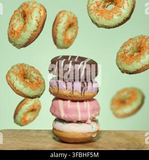 A stack of four brightly glazed donuts on a table with green donuts with orange sprinkles flying around them Stock Photo