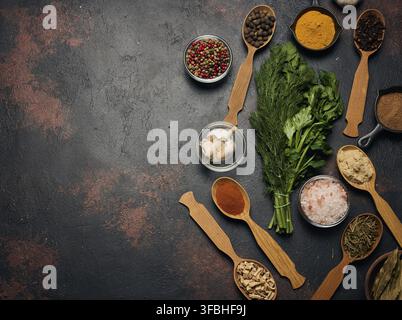 Spices, herbs, and garlic on a dark background. Turmeric, paprika, pepper, cloves, ginger, and salt in bowls and spoons. Fresh dill and parsley. Top v Stock Photo