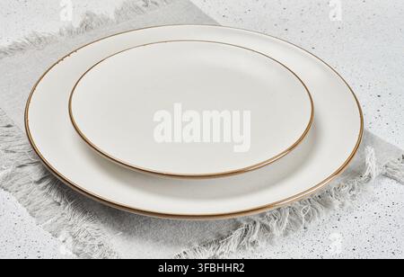 Two empty white ceramic plates with brown rims stacked on a gray linen napkin with fringe Stock Photo
