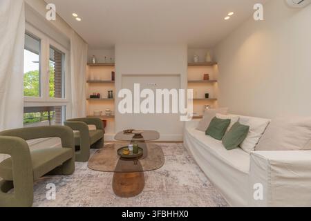 Cozy neutral living room with olive green armchairs, glass coffee tables, built-in shelving, and white sofa Stock Photo