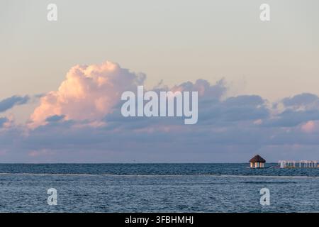 Calm blue ocean beneath soft pink sunset clouds, with a small thatched hut on the horizon and distant coastal buildings. Stock Photo