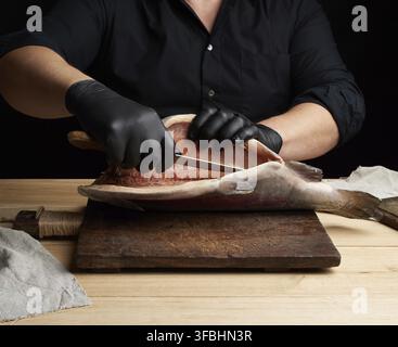 Chef in black uniform and black latex gloves cuts a carcass of fresh salmon fish on a wooden cutting board, black background Stock Photo
