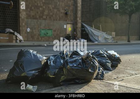 Black garbage bags lie on the edge of a city street near a homeless shelter made from tarps and cardboard. The gritty urban background shows poverty, Stock Photo