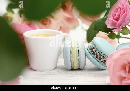 White ceramic cup with coffee and macarons on a white table, behind a bouquet of pink roses, close up Stock Photo